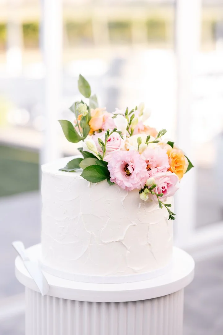 A white frosted cake decorated with pink, white, and peach flowers and green leaves, set on a round white cake stand.