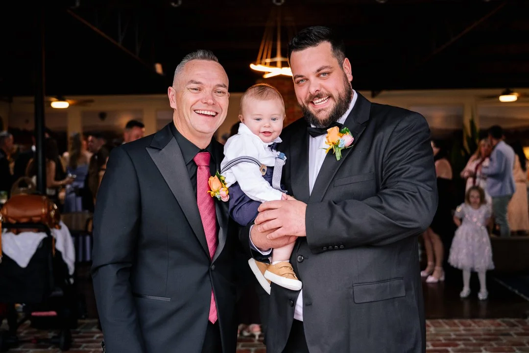 Two men in suits and a young child dressed up at a celebration or social gathering with other people in the background.