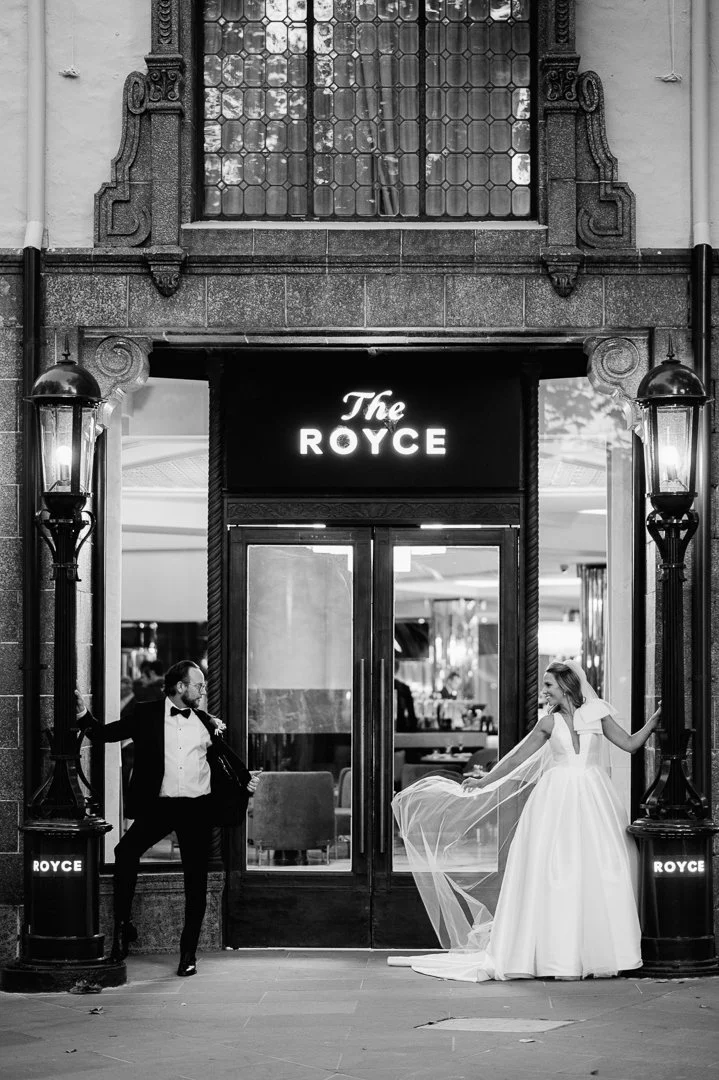 Black and white photo of a man in a tuxedo and a woman in a wedding dress posing outside the entrance of The Royce, with vintage-style lanterns on either side of the doorway.