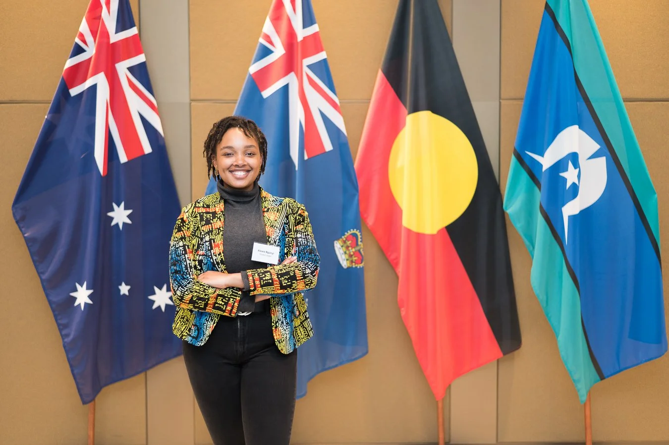 A smiling woman stands with arms crossed in front of four flags, including the Australian, New Zealand, Aboriginal, and Torres Strait Islander flags.