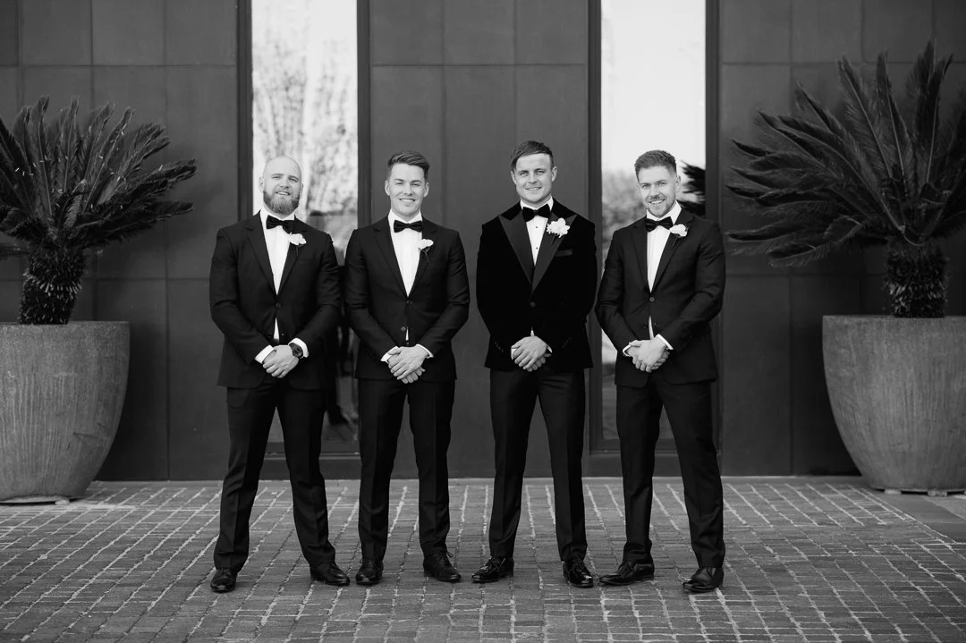 Four men in tuxedos and bow ties standing outdoors in front of a building with large potted plants on each side.