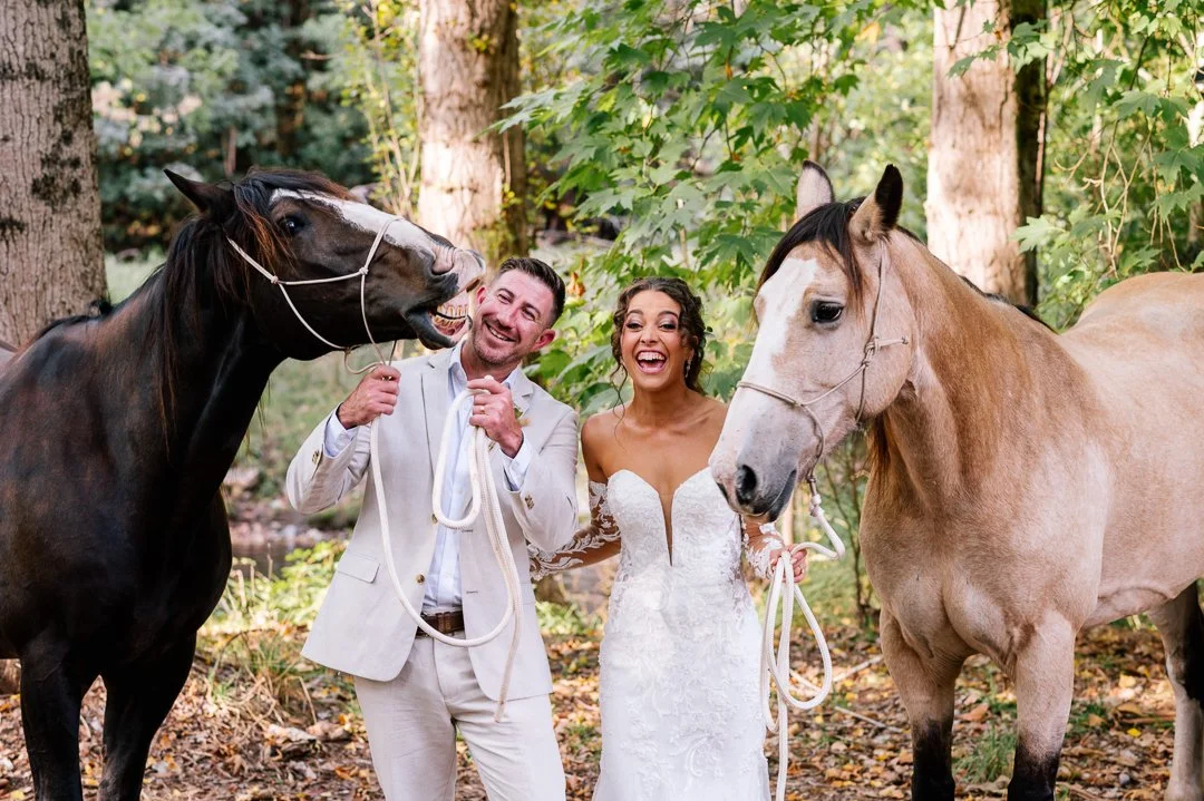 A newlywed couple in wedding attire in a forest, holding horses' reins and smiling.