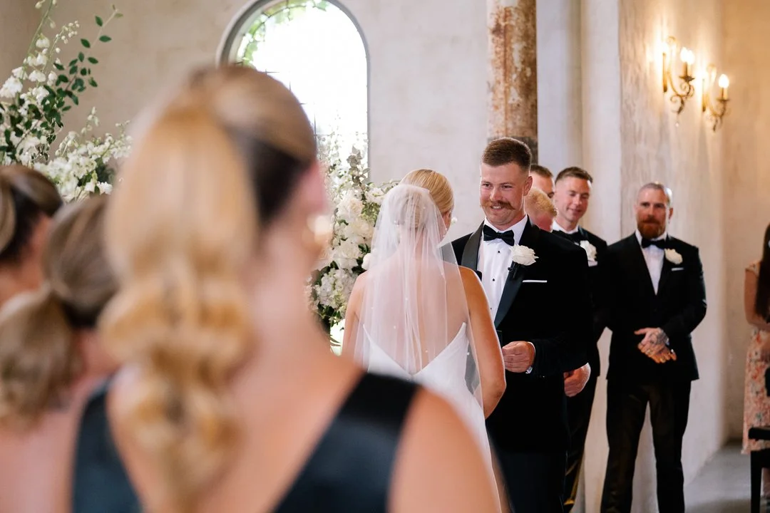 A wedding ceremony with a bride and groom exchanging vows, surrounded by bridesmaids and groomsmen in formal attire, inside a decorated venue with floral arrangements and soft lighting.