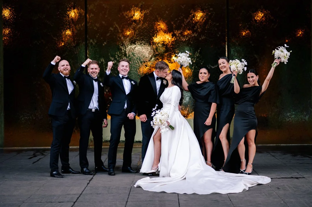 A wedding party with seven people, including a bride and groom in the center, surrounded by four bridesmaids and two groomsmen. The bride holds a bouquet, and the groom leans in to kiss her. The groomsmen are dressed in tuxedos, and the bridesmaids a