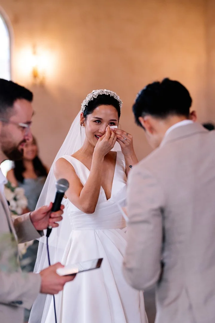 A bride in a white wedding gown and veil, holding a tissue to her face, smiling during her wedding ceremony, with an officiant holding a microphone and reading from a paper, and a groom in a light-colored suit standing opposite her.