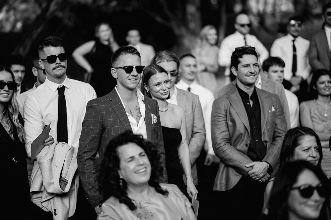 A black-and-white photo of a group of people at an outdoor event, with some wearing sunglasses and formal attire, listening to a speaker or watching a performance.