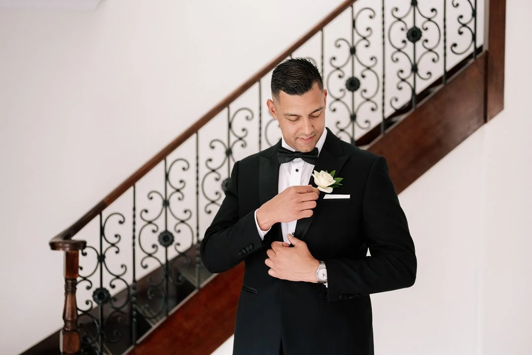 Man in black tuxedo with bow tie adjusting a white boutonnière on his suit, standing in front of a staircase with ornate black iron railing.