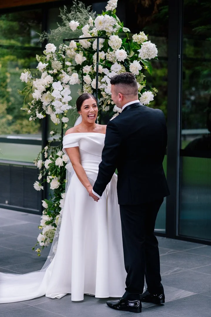 A bride and groom holding hands during their wedding ceremony, smiling at each other, with a floral backdrop of white flowers and greenery behind them.