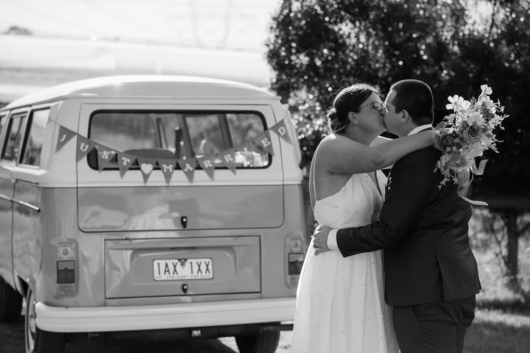 A bride and groom kissing in front of a vintage van with a 'Just Married' banner, outdoor setting, black and white photo.