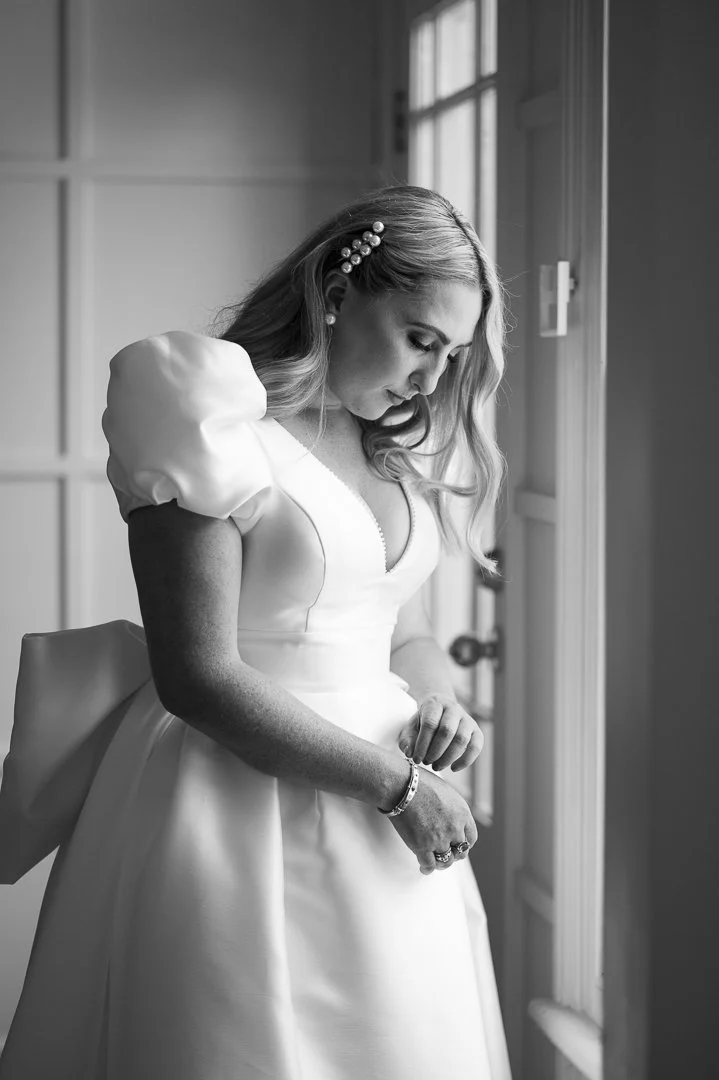 A woman in a white wedding dress, standing by a window, looking down thoughtfully, with her hair styled with pearl hair clips, in black and white.