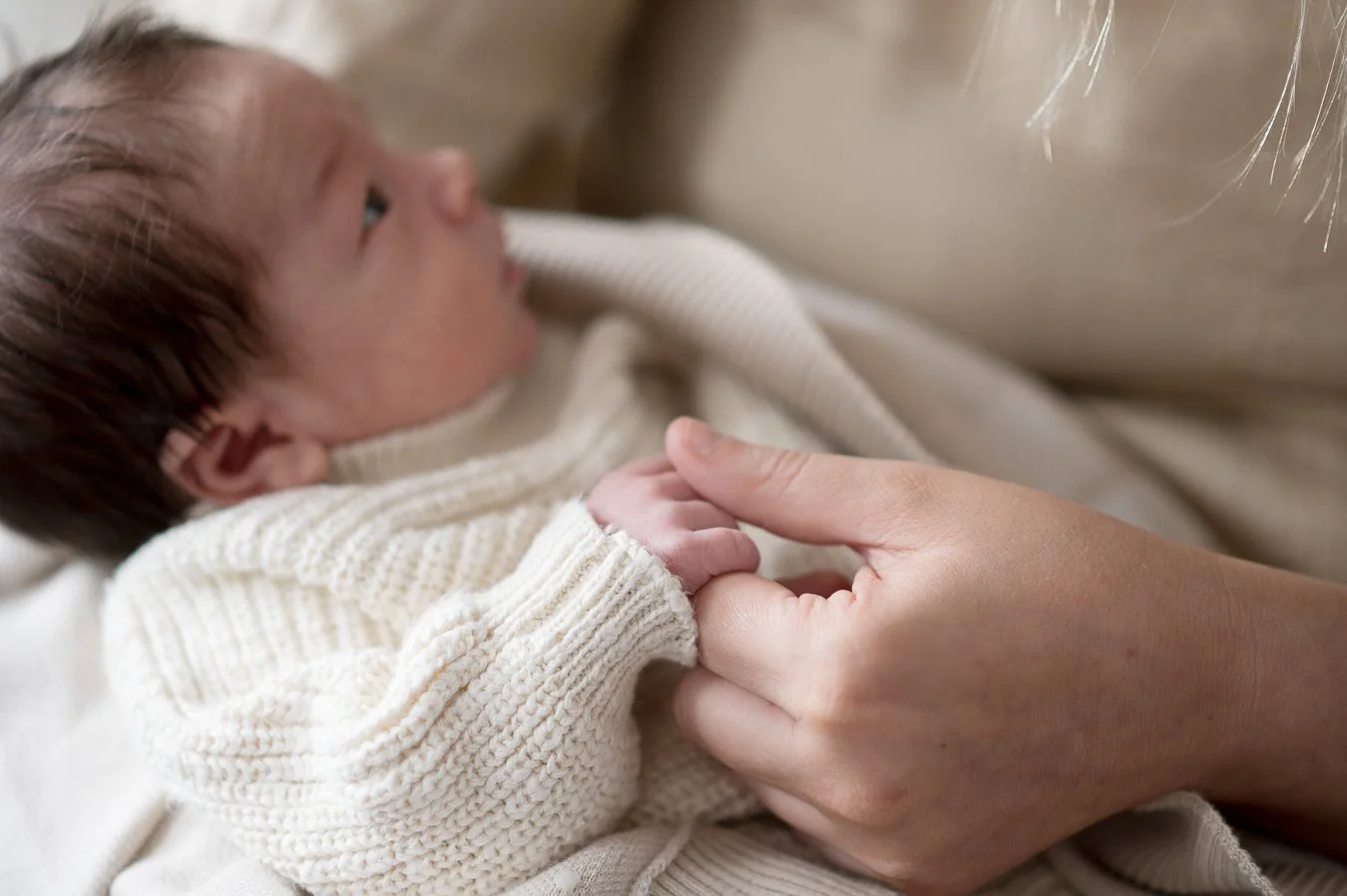 A newborn baby lying on a blanket, gently held by an adult hand, wearing a cream-colored knitted sweater.