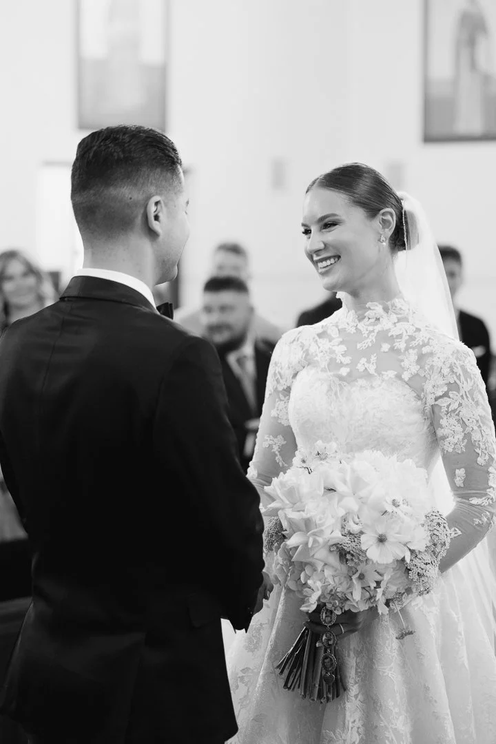 Black and white photo of a bride and groom at their wedding ceremony, with the bride smiling and holding a bouquet of flowers, and the groom facing her, in a room with guests in the background.