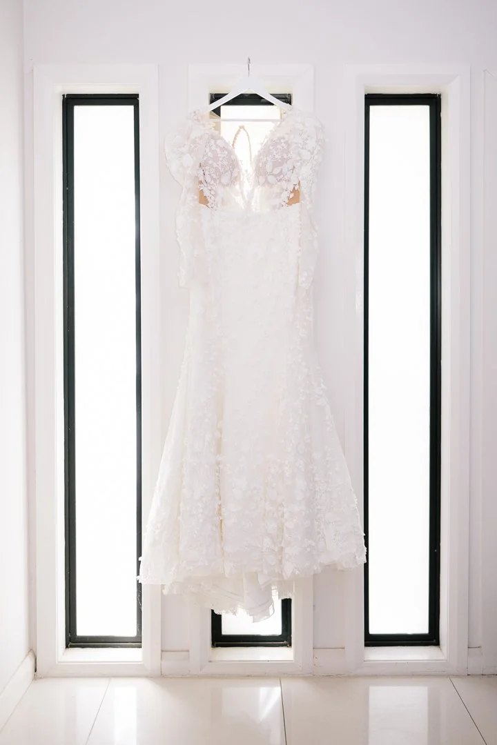 A white lace wedding dress hanging on a white hanger in front of three windows.