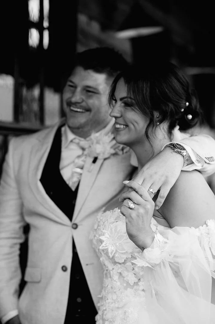 Black and white photo of a smiling bride and groom on their wedding day, with the groom's arm around the bride's shoulder. The bride is showing her wedding ring and wearing a lace wedding dress. Both appear happy and joyful.