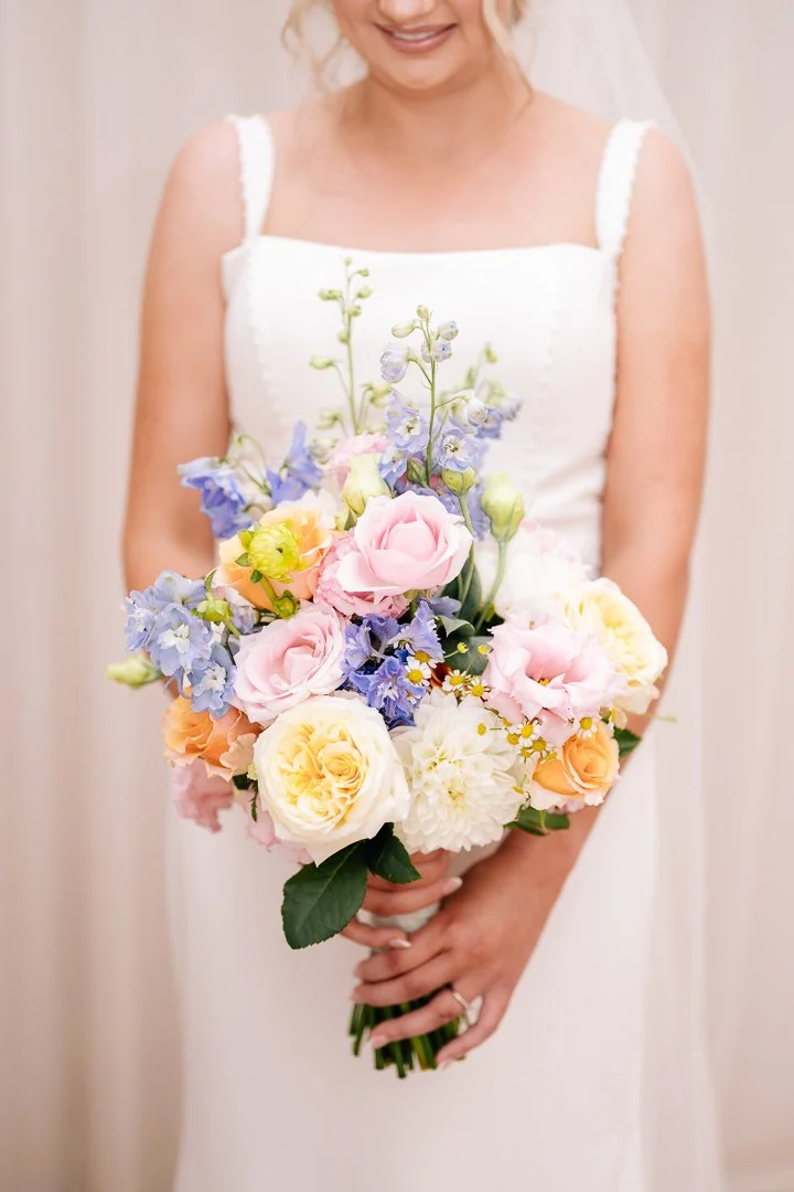 A woman in a white dress holding a bouquet of pastel-colored flowers including roses, daisies, and delphiniums.