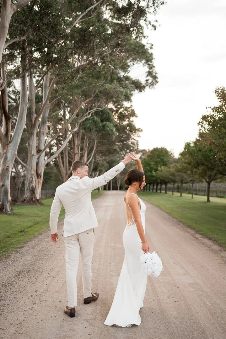 A newlywed couple dancing and holding hands on a dirt path lined with trees, with the groom twirling the bride.