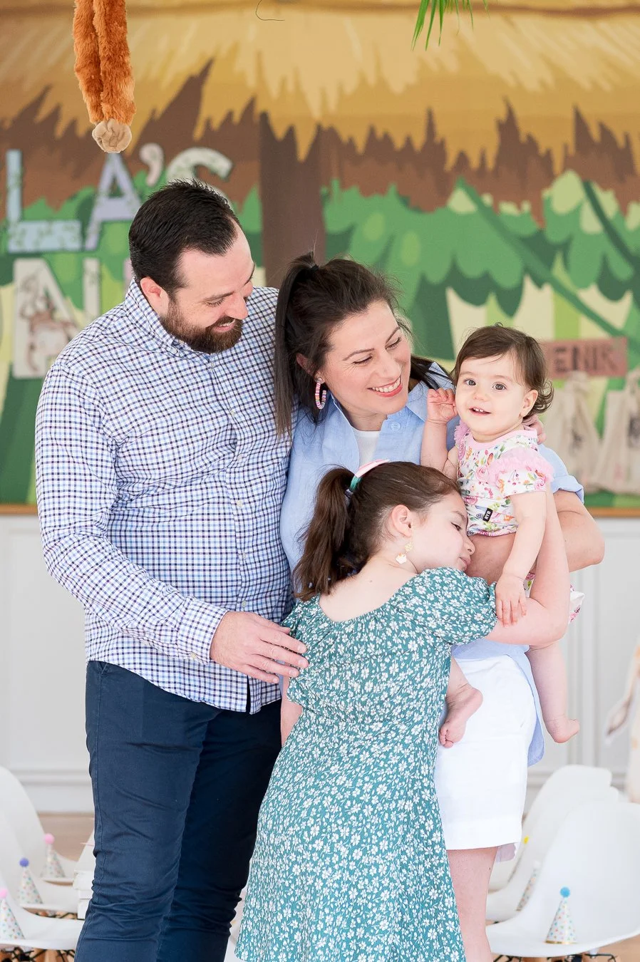 A happy family of four, including a man, a woman, and two young girls, sharing a joyful moment together in a colorful, decorated room.
