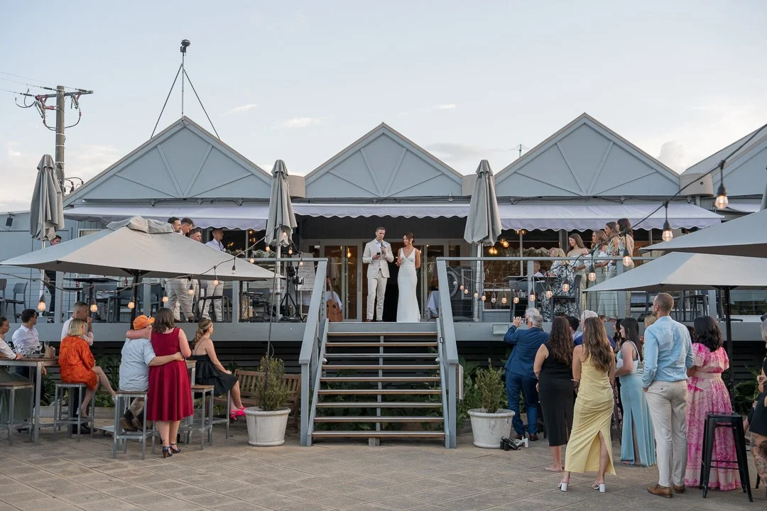 Wedding celebration with bride and groom on an outdoor deck, surrounded by guests, during the evening with string lights and umbrellas.