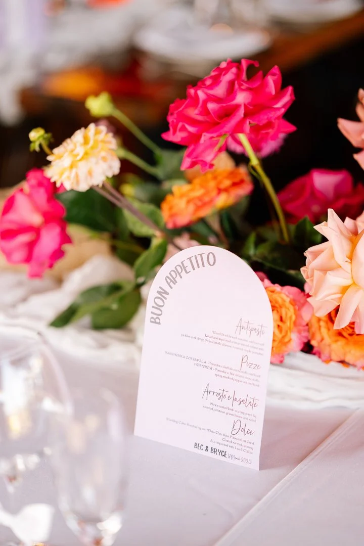 A colorful bouquet of pink, peach, and orange flowers with green leaves, placed on a table next to a white menu card with Italian food options.