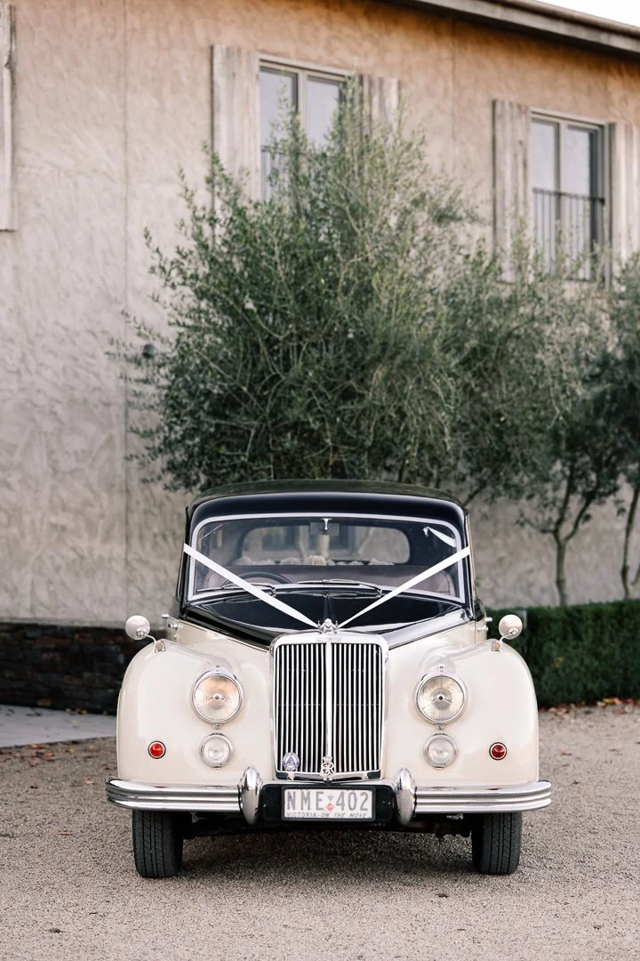 A vintage white car with a black roof, decorated with white ribbon, parked on a gravel driveway in front of a house with beige walls and two windows with shutters.