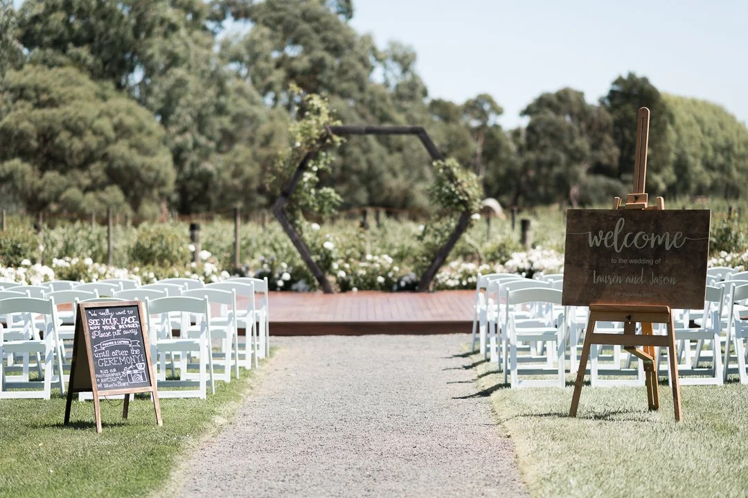 Outdoor wedding setup with white chairs arranged on either side of a grassy aisle leading to a decorated hexagonal arch and a welcome sign for Lauren and Jason.