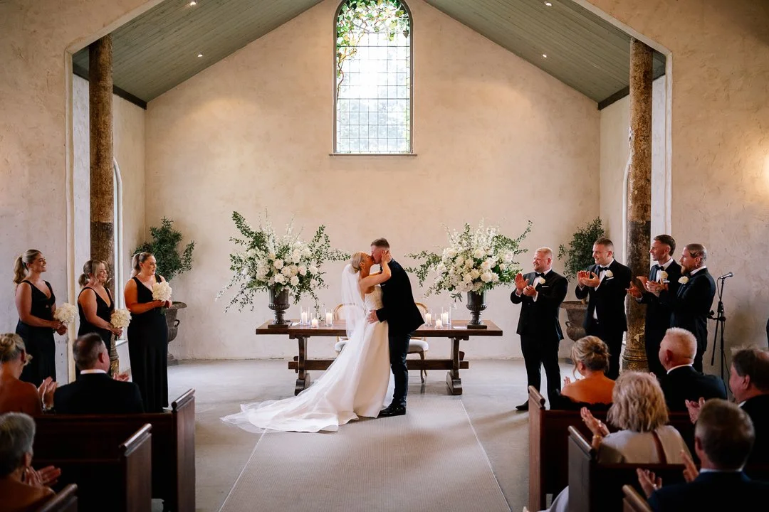 A wedding ceremony in a church with a bride and groom kissing at the altar, surrounded by bridesmaids and groomsmen clapping, and guests seated watching.