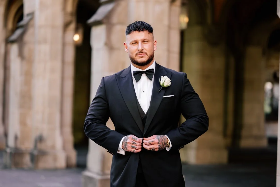 A man wearing a black tuxedo with a bow tie and a white boutonniere, standing outdoors in front of stone arches.