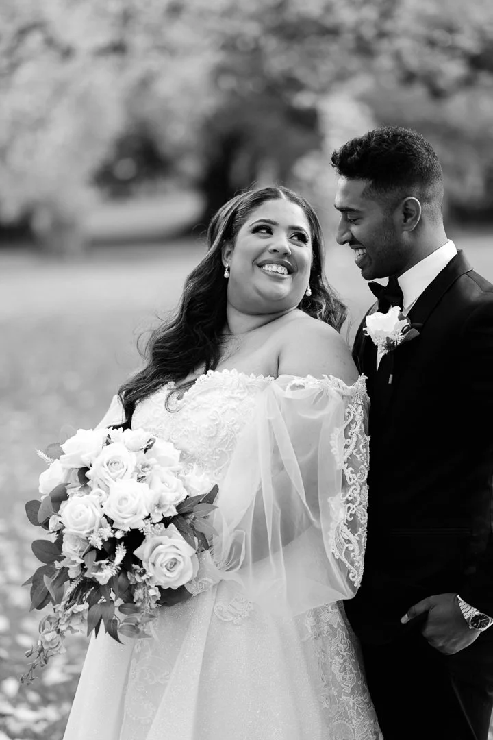 Black and white photo of a bride and groom standing outdoors, the bride holding a bouquet of roses and smiling at the groom, who is leaning toward her in a tuxedo.