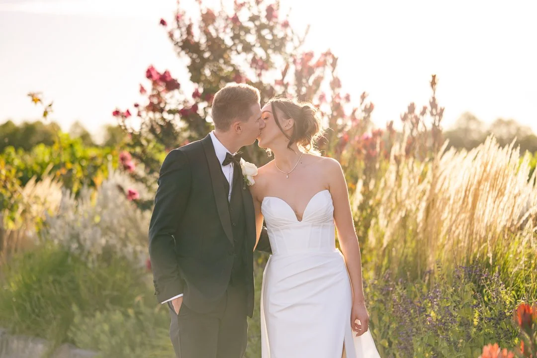 A newlywed couple sharing a kiss outdoors in a garden during sunset, with the bride in a white strapless wedding gown and the groom in a black tuxedo.