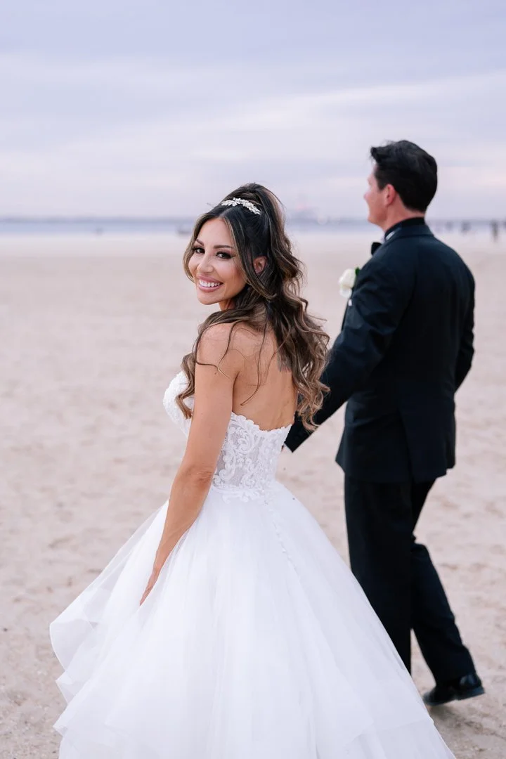 A bride and groom holding hands on the beach, with the bride smiling at the camera. The bride is wearing a white strapless wedding gown with lace details, and the groom is in a black tuxedo. The beach and ocean are visible in the background.