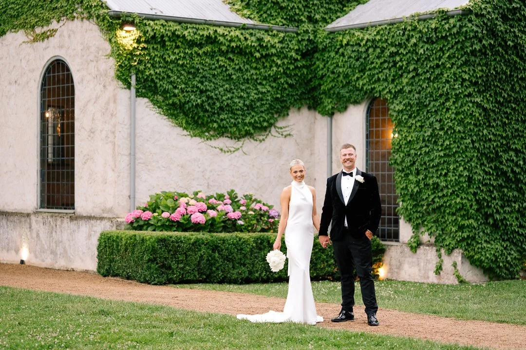 A bride and groom standing on a grass pathway outside a building covered with green ivy. The bride is in a white wedding dress holding a bouquet, and the groom is in a tuxedo with a bow tie, both smiling.