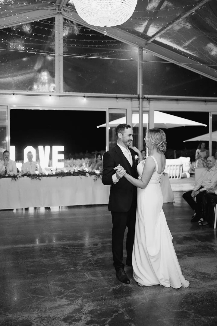 A couple is dancing at their wedding reception inside a decorated venue with string lights and a large illuminated 'LOVE' sign in the background. The groom is in a suit, and the bride is in a wedding gown. Guests are seated at tables around them.