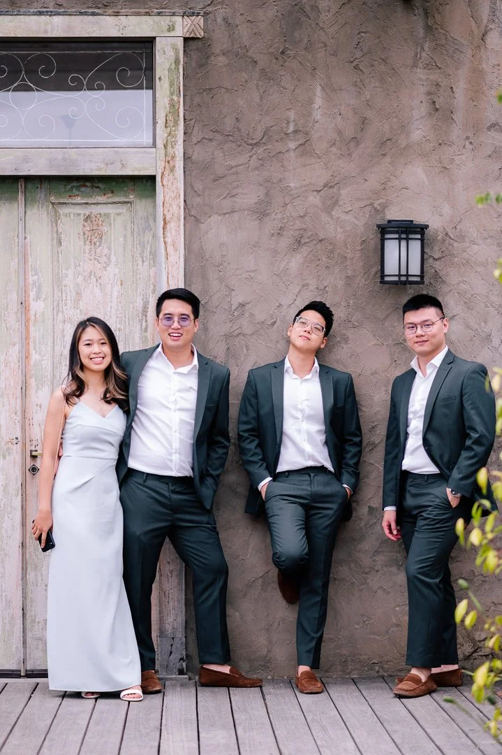 Four young adults dressed in formal attire, standing outdoors against a rustic wall, smiling and posing for the photo.