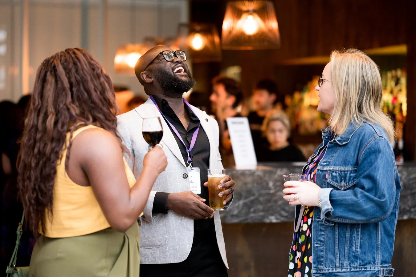 Three people socializing at a bar, two women and one man, holding drinks and laughing, with a bar counter and other patrons in the background.