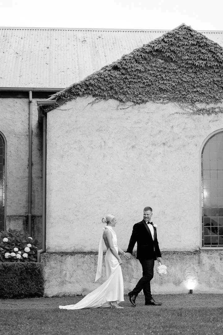A bride and groom holding hands and walking outside a building with large windows, in black and white.
