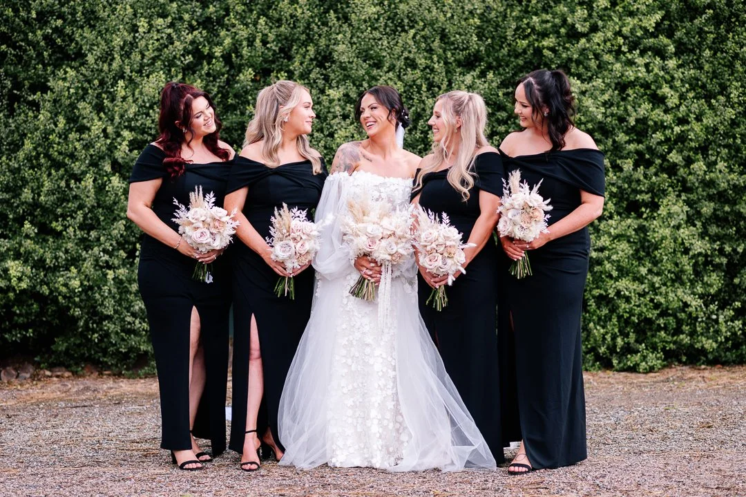 A bride and four bridesmaids standing outdoors against a green hedge, all holding bouquets of flowers and smiling.