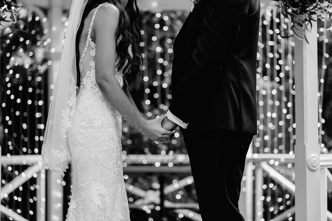 A bride and groom holding hands during their wedding ceremony with string lights in the background, in black and white.