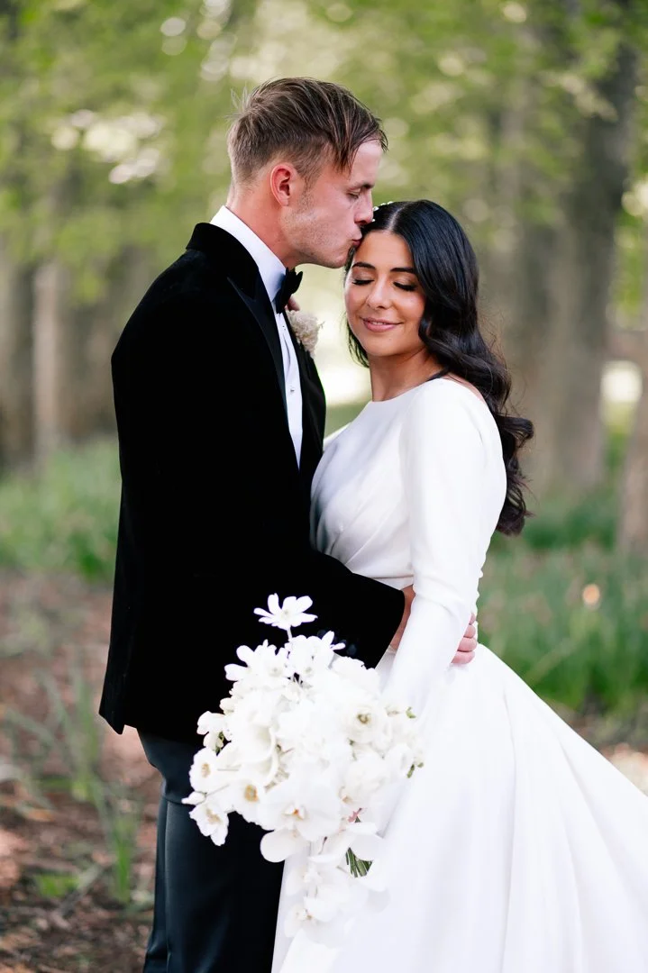 Bride and groom embracing outdoors, groom kissing bride's forehead, bride holding a white flower bouquet, both dressed in wedding attire with a blurred green forest background.