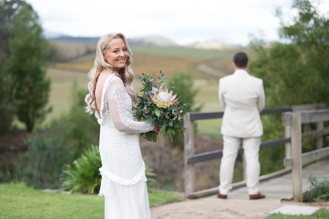 A woman in a white dress holding a bouquet of flowers, smiling, with a man in a white suit standing on a bridge and looking away in an outdoor setting.