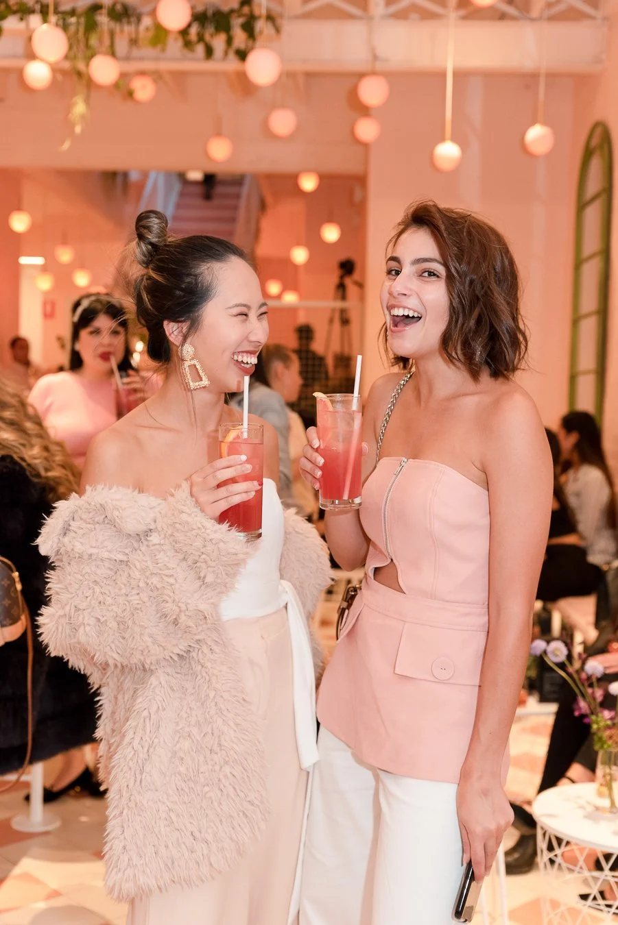 Two women smiling and laughing while holding pink drinks at a social gathering or party, with warm lighting and other people visible in the background.