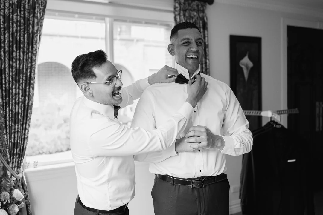 Two men helping each other put on tuxedo bow ties in a well-lit room with floral curtains and artwork on the walls.