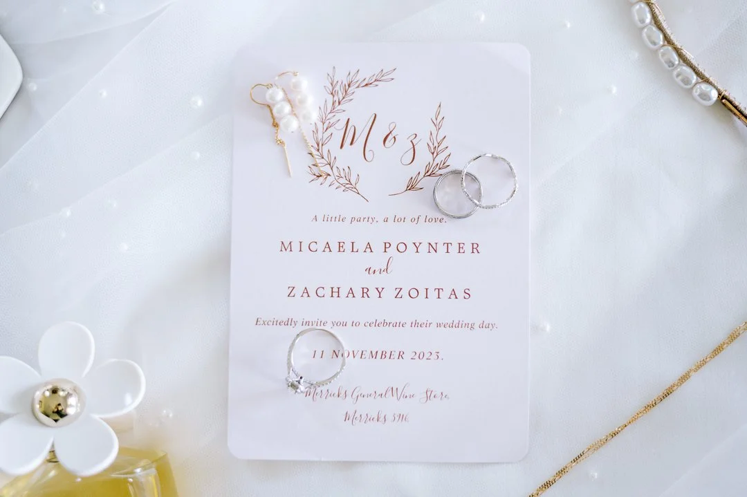 Wedding invitation card with two rings, a pearl hair accessory, and jewelry items on a white fabric background.