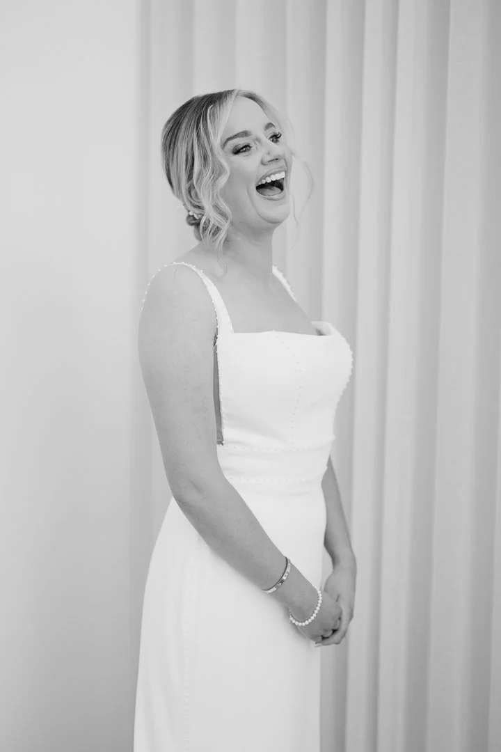 A woman in a white dress smiling and laughing, standing indoors with curtains in the background.