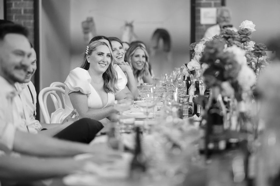 A group of people at a formal event, sitting at a long table decorated with flowers and drinks, smiling and engaging in conversation.