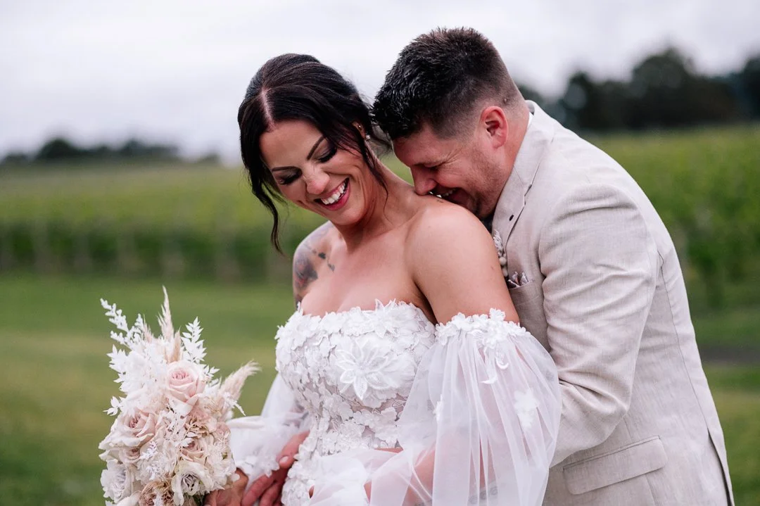 A smiling woman in a white wedding dress holding a bouquet of flowers, with a man in a light-colored suit leaning close to her, outdoors in a grassy area with a blurred green landscape in the background.