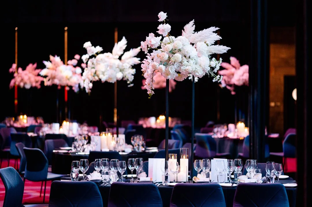 Elegant event setup with tall floral centerpieces featuring white and pink flowers on round tables, dim lighting, and candlelit ambiance.