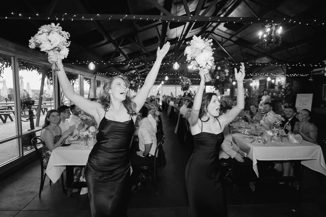 Two women in black dresses celebrating, holding bouquets, with guests sitting at decorated tables in the background at a wedding reception.