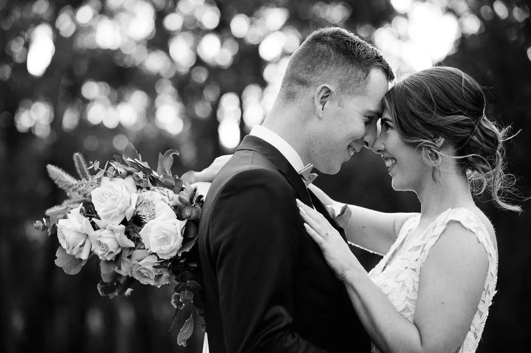 A black and white photograph of a bride and groom embracing outdoors, leaning their foreheads together and smiling, with a bouquet of flowers held behind the groom.