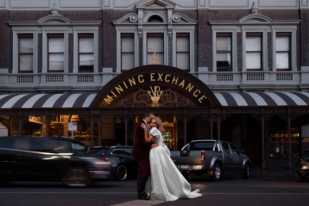 Couple in wedding attire sharing a romantic moment in front of a historic building with a sign that reads 'Mining Exchange' and several parked cars on the street.