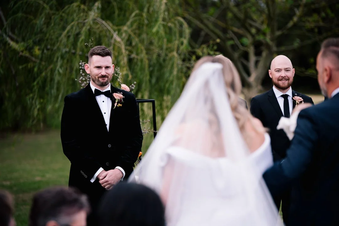 Groom and officiant at outdoor wedding ceremony, with bride and guests partially visible